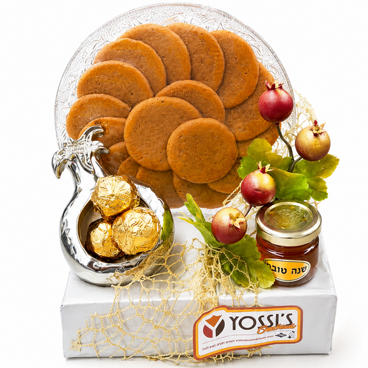 A gift basket of cookies and treats from Yossi's, featuring a large plate of round golden-brown cookies, a silver rooster-shaped container filled with gold-wrapped chocolates, and a small jar of honey, arranged on a white box with decorative netting and pomegranate branches on a white background.