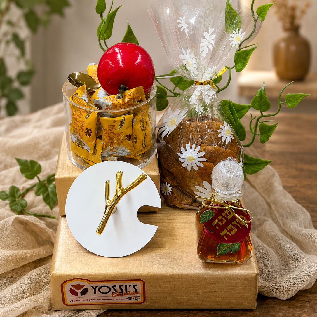 A gift set arranged on a wooden box branded Yossi's, featuring a small jar of yellow candies topped with a red apple, a wrapped brown loaf with white daisy print and green leaves, a small amber bottle, and a white round tray with a gold branch-shaped ornament, all styled on beige fabric.